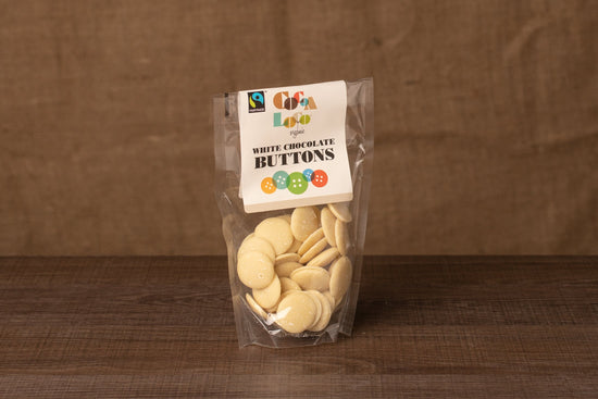 A packet of white chocolate buttons, with the Cocoa Loco label, on a dark wood counter with a brown background